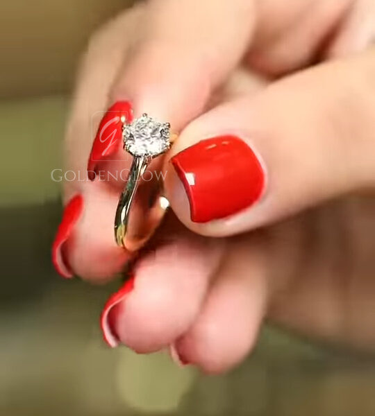 A top-down angled perspective of the ring on the finger. The round brilliant diamond or Moissanite is the star, showcasing a crisp "arrows" facet pattern indicative of an ideal cut. The yellow gold prongs are visible at the corners, securely holding the gem. The band blurs into the background, focusing attention on the stone's clarity and lack of inclusions. This shot demonstrates the premium quality of the eco-friendly gemstone and its colorless brilliance.