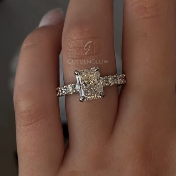 A captivating front-facing view of a massive elongated radiant-cut solitaire engagement ring held between fingers. The centerpiece is a premium lab grown diamond or Moissanite, showcasing a "crushed ice" facet pattern that refracts light into a dazzling display of sparkles. The stone is secured by four claw prongs in polished 18k yellow gold. This image highlights the stone's impressive size and the timeless elegance of the minimalist setting that allows the gem to be the absolute focus.