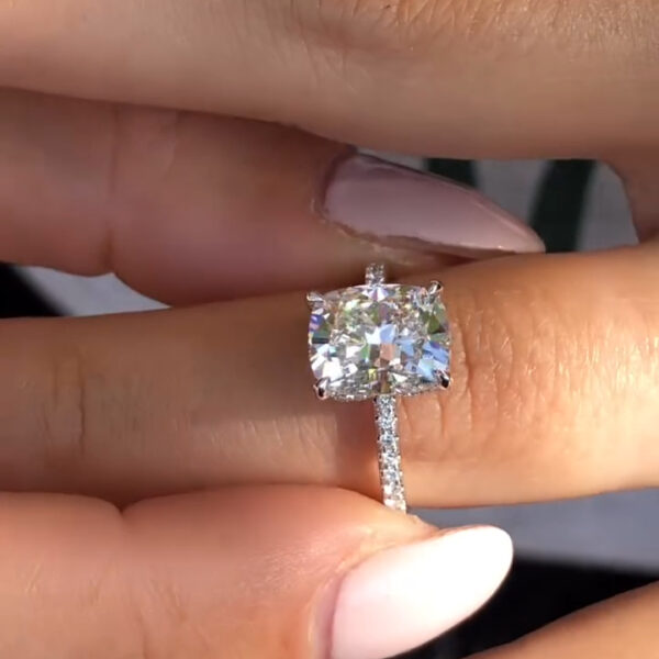 A detailed side-profile view of the ring held vertically between fingers. This angle reveals the structural beauty of the "hidden halo" design—a row of tiny diamonds or Moissanites wrapping around the gallery rail beneath the main stone. The elongated cushion-cut lab grown diamond is secured by four claw prongs. The pavé-set band glitters even from the side. This shot emphasizes the ring's depth and the intricate hidden touches that make it a bespoke luxury piece.