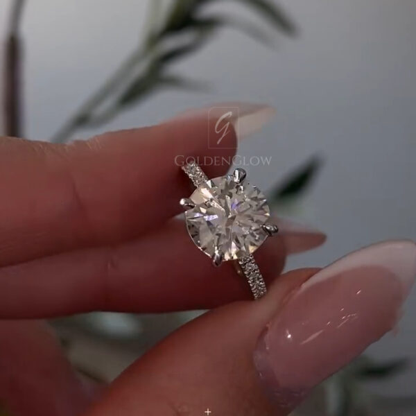 A stunning side-profile view of a handcrafted oval halo engagement ring held between fingertips. This angle reveals the sophisticated architecture of the setting, where the main oval-cut lab grown diamond or Moissanite rests high in a basket. The image highlights the "cathedral" style shank that rises to meet the halo, adding structural elegance. The band is lined with micro-pavé stones that glitter even from the side. You can clearly see the depth of the center stone and the delicate metalwork supporting the halo.