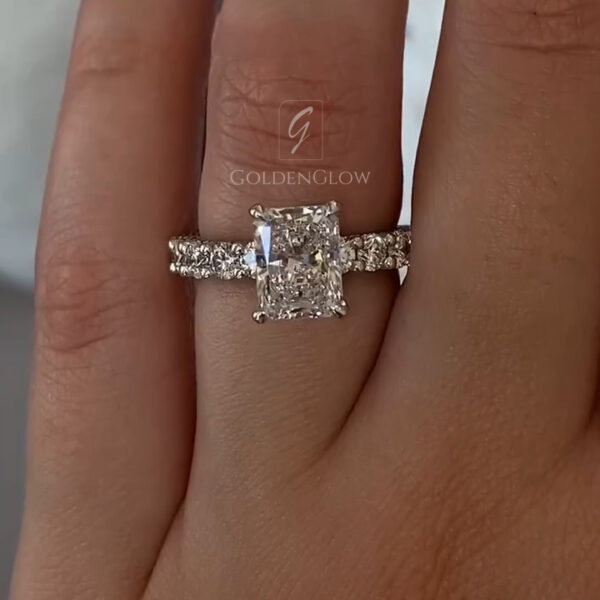 Close-up view of a radiant-cut engagement ring worn on the finger, featuring a sparkling diamond set in a classic four-prong setting. The elongated radiant shape reflects light beautifully, creating crisp brilliance across the stone. A pavé-set band lined with round accent stones adds extra shimmer and luxury. Crafted for modern elegance, this engagement ring is also available with a moissanite diamond, offering a sustainable and ethical alternative with exceptional fire and durability.