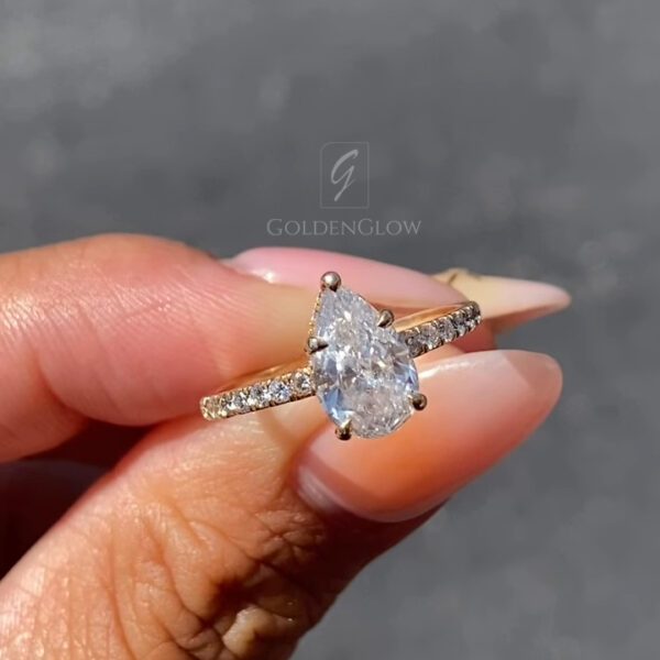 Front view of a pear-shaped solitaire engagement ring held between fingers in bright natural light. The center diamond is secured in delicate prongs and sparkles with crisp brilliance, while a pavé-set band adds subtle shimmer along the rose gold shank. Elegant and timeless, this pear-cut engagement ring is designed for modern romance and is also available with a moissanite diamond option, offering an ethical, sustainable alternative with exceptional fire and clarity.