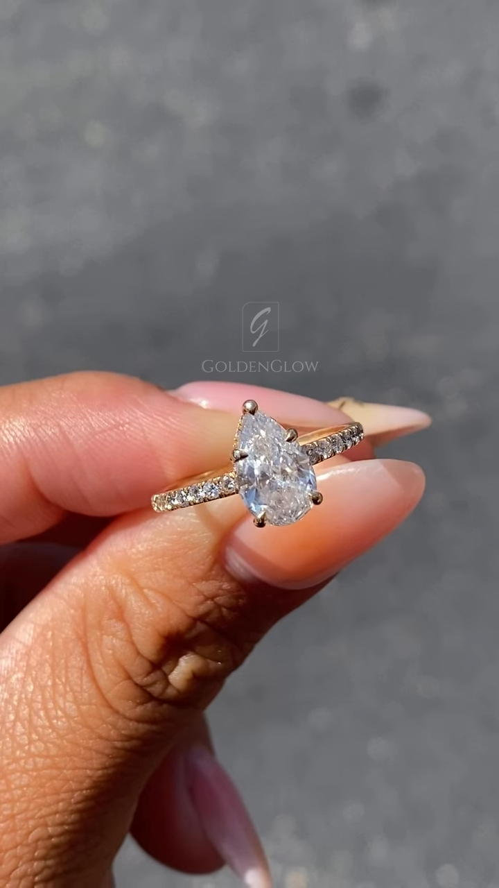 Front view of a pear-shaped solitaire engagement ring held between fingers in bright natural light. The center diamond is secured in delicate prongs and sparkles with crisp brilliance, while a pavé-set band adds subtle shimmer along the rose gold shank. Elegant and timeless, this pear-cut engagement ring is designed for modern romance and is also available with a moissanite diamond option, offering an ethical, sustainable alternative with exceptional fire and clarity.