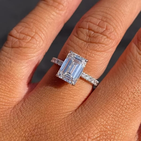Angled close-up of an emerald-cut solitaire engagement ring held between fingers against a soft gray background. The diamond showcases sharp corners and elongated proportions, while the pavé band adds subtle shimmer along the shank. Designed for timeless elegance and everyday wear, this engagement ring can also be customized with a moissanite diamond, offering a sustainable alternative with radiant sparkle and durability.