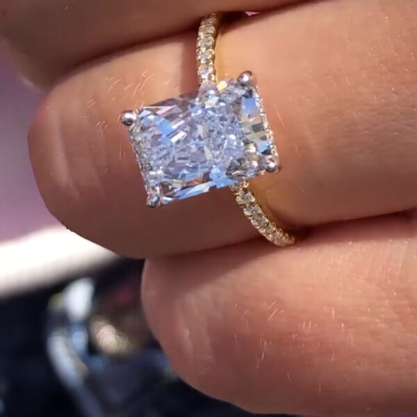 Close-up side view of a rectangular engagement ring worn on a hand, featuring a brilliant diamond secured in a four-prong setting with a slim yellow gold pavé band. The stone reflects bright flashes of light, highlighting crisp facets and clean lines. This image compares the refined sparkle and structure of a lab grown diamond to the rainbow fire often associated with a moissanite diamond, shown in natural outdoor lighting.
