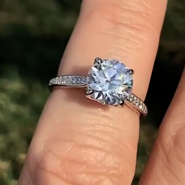 Angled front view of a round diamond engagement ring on the hand, showcasing a diamond with strong white sparkle and symmetrical facets. The pavé-accented band enhances the center stone without overpowering it. Outdoor lighting creates visible flashes of light. This photo helps viewers compare the refined brilliance of a lab grown diamond with the rainbow fire typically seen in a moissanite diamond.