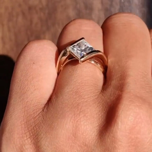 Top-down view of a princess-cut solitaire engagement ring on the hand, featuring a sleek yellow gold band with a contemporary bypass setting. The center stone catches sunlight, revealing sharp facet definition and mirror-like reflections. The image highlights the refined beauty and fire seen in both a lab grown diamond and moissanite diamond, offering a clear comparison of brilliance, cut precision, and modern minimalist ring styling.