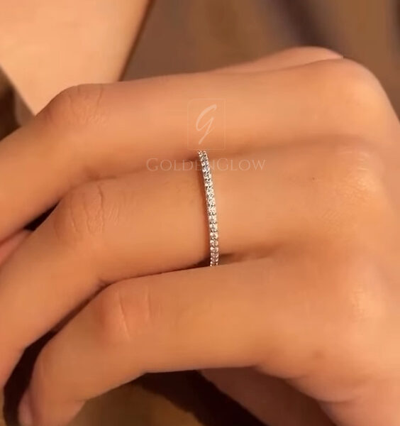 Angled view of a thin diamond band on a finger, showcasing the uniform line of round-cut diamonds set along the band. The warm lighting enhances the brilliance of the stones while maintaining a soft, romantic tone. The ring appears lightweight and delicate, ideal for stacking or wearing alone. The close framing focuses on the diamond detail and smooth metal finish, with the background kept neutral to emphasize the jewelry.