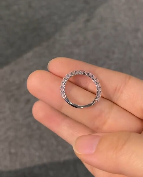 Angled perspective of a delicate eternity band pinched gently between thumb and forefinger, revealing the side profile of the setting. The stones are uniform in size and mounted in a continuous pattern around the ring, allowing light to pass through the gallery beneath. The neutral gray background keeps focus on the brilliance and symmetry, making the ring suitable for comparison between a lab grown diamond and a moissanite diamond design.