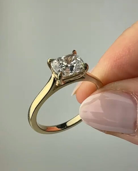 A minimalist yellow gold ring is displayed between fingertips, focusing on the emerald-cut center stone raised in a classic prong setting. The gemstone catches light across its clean facets, emphasizing symmetry and brilliance. The smooth gold band curves gently, offering a timeless look. This image is ideal for comparing a lab grown diamond and a moissanite diamond in a refined solitaire engagement ring design.