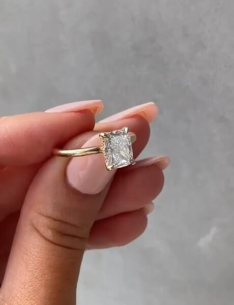 Side-angle view of a rectangular solitaire ring in yellow gold, photographed in natural light while held by hand. The elevated basket setting reveals the depth and structure of the center stone, with clean prongs and a smooth, narrow band. The gemstone reflects sharp flashes as it tilts, making this image useful for visual comparison between a lab grown diamond and a moissanite diamond in modern engagement ring design.