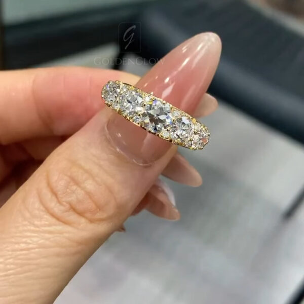 Close-up of a yellow gold diamond band held delicately between manicured fingers with glossy nude-pink nails. The ring is shown from a narrow side angle, revealing a gentle taper in the band and a row of round brilliant diamonds set closely together. The diamonds catch soft ambient light, creating subtle flashes. The background is softly blurred, suggesting an indoor setting with neutral tones, allowing the gold and diamonds to remain the focal point.