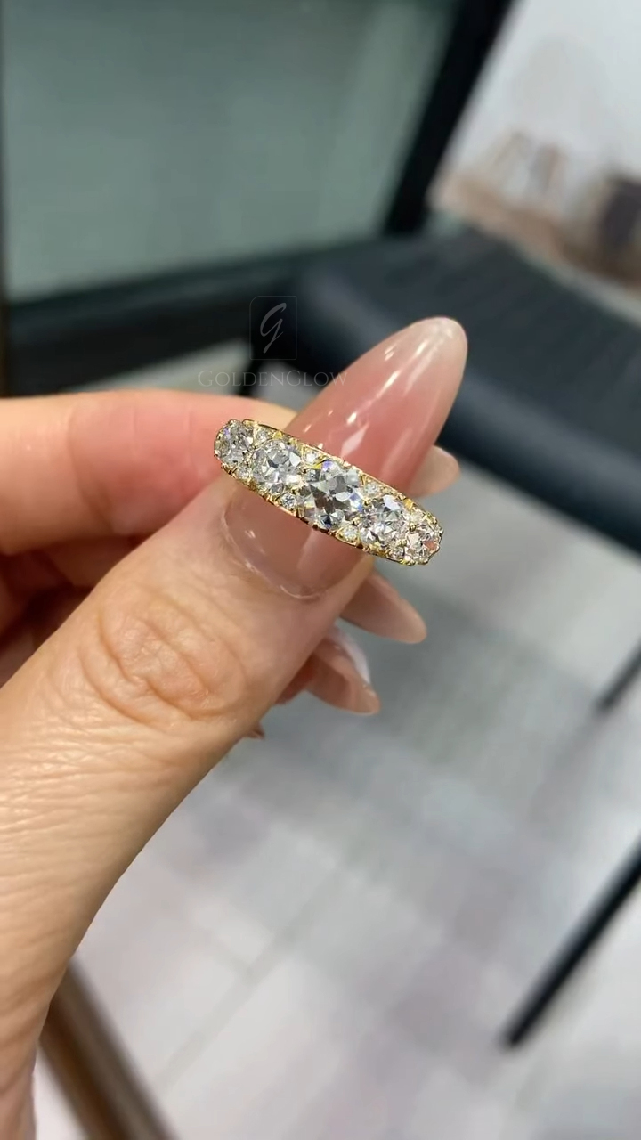Close-up of a yellow gold diamond band held delicately between manicured fingers with glossy nude-pink nails. The ring is shown from a narrow side angle, revealing a gentle taper in the band and a row of round brilliant diamonds set closely together. The diamonds catch soft ambient light, creating subtle flashes. The background is softly blurred, suggesting an indoor setting with neutral tones, allowing the gold and diamonds to remain the focal point.
