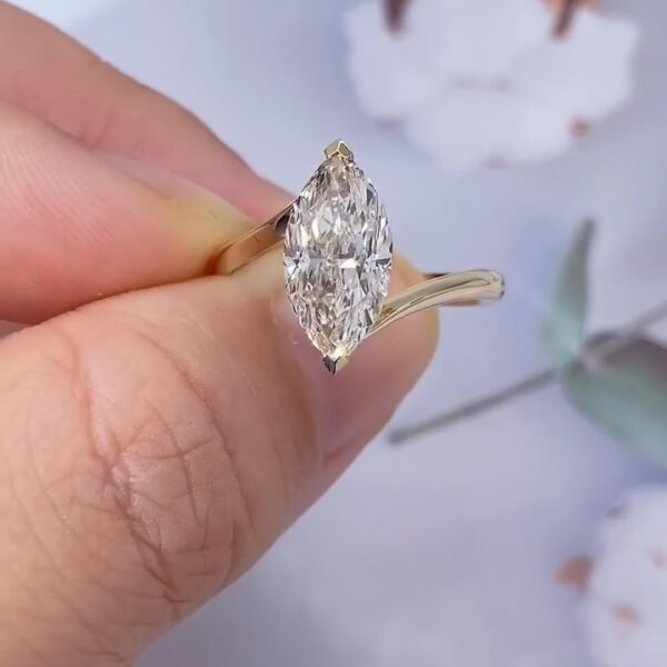 The same marquise diamond ring is shown from a slightly adjusted angle, revealing more of the stone’s depth and internal reflections. The yellow gold band curves smoothly toward the setting, creating a flowing silhouette. Light glints across the diamond’s pointed tips, highlighting the symmetry of the cut. The soft-focus floral background remains visible, adding a delicate, romantic atmosphere without distracting from the ring’s craftsmanship.