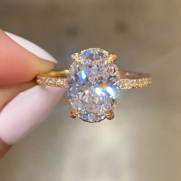 Close-up of an oval diamond or moissanite engagement ring in yellow gold, held between manicured fingers. The oval brilliant center stone sparkles with rainbow fire, secured by four claw prongs. A delicate pavé band adds subtle shimmer, while the softly blurred background highlights the stone’s clarity and brilliance.