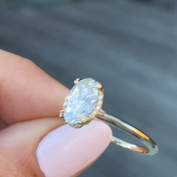 Angled profile shot of an oval-cut diamond or moissanite ring resting against a softly blurred urban background. The oval gemstone displays exceptional fire and clarity, framed by sculpted prongs and a refined hidden halo. The yellow gold band is slender and minimalist, allowing the center stone to remain the focal point. This image emphasizes ethical luxury, modern design, and elegance.