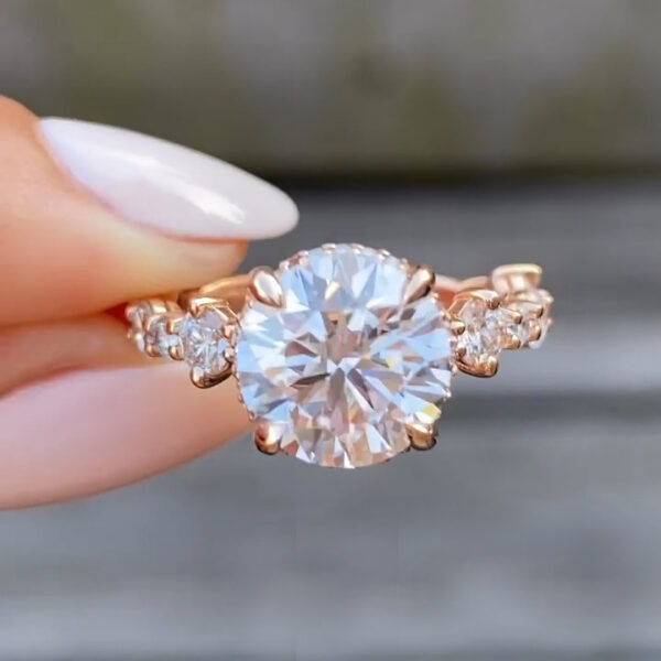 Close-up side view of a diamond or moissanite engagement ring in warm rose gold, held between manicured fingers. The oval brilliant-cut center stone sparkles with strong fire and clarity, secured by sculpted prongs. Pavé accent stones line the band, adding shimmer along the shoulders. The polished rose gold setting reflects light softly, highlighting ethical craftsmanship, sustainable materials, and fine jewelry detail suitable for modern bridal and luxury engagement ring collections.
