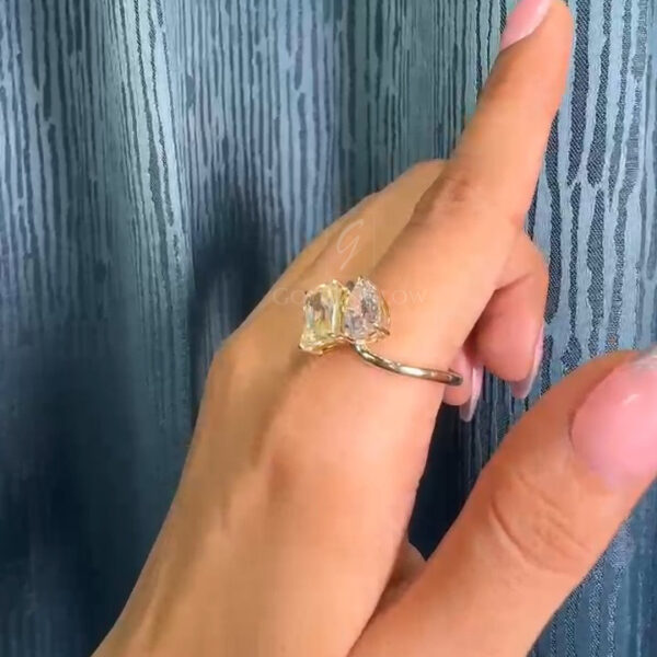 Side angle view of a diamond and moissanite toi et moi ring on a hand, showing the emerald-cut stone and pear-shaped accent set in polished yellow gold. The slim band and elevated setting emphasize the modern, asymmetrical design and light-catching facets.