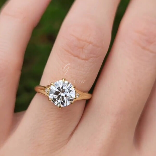 Close-up view of a yellow gold engagement ring worn on a finger outdoors. The ring features a round brilliant-cut diamond set in a classic prong setting, catching natural light and reflecting bright flashes. The warm gold band contrasts with the cool sparkle of the stone. Soft green foliage in the background is blurred, creating a natural, romantic setting that highlights the diamond’s brilliance and the elegant proportions of the ring.