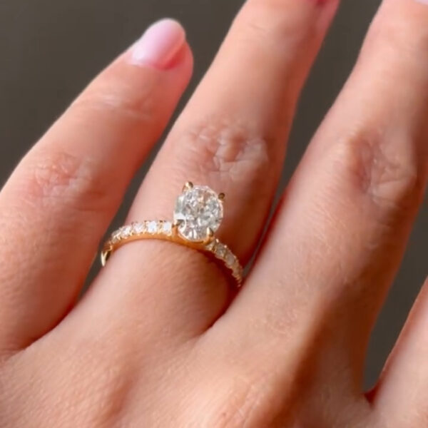 A hand wearing a yellow gold engagement ring with an oval-cut diamond center stone. The diamond is held by four prongs and sits on a delicate pavé diamond band. The ring is photographed at an angle on the finger, showing how the stone catches light and reflects brilliance while maintaining an elegant, minimal profile.