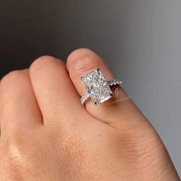 Angled top view of a radiant-cut diamond ring resting on a finger, showcasing the stone’s faceting and crisp corners. The center diamond catches light across its table and crown, producing bright flashes. The pavé diamond band enhances brilliance while remaining delicate and refined. Natural skin texture and soft lighting create a realistic presentation, emphasizing craftsmanship, sparkle, and the luxurious appearance of the engagement ring.