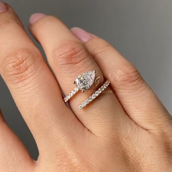 Close-up view of a pear-shaped diamond wrap ring worn on a hand, featuring a brilliant pear-cut center stone set in warm rose gold. The open bypass design curves gracefully around the finger, with pavé-set diamonds lining both arms of the band. The center diamond catches the light, creating sharp flashes and soft sparkle, while the slim diamond-studded band adds elegance. Neutral gray background highlights the refined craftsmanship and modern bridal style.