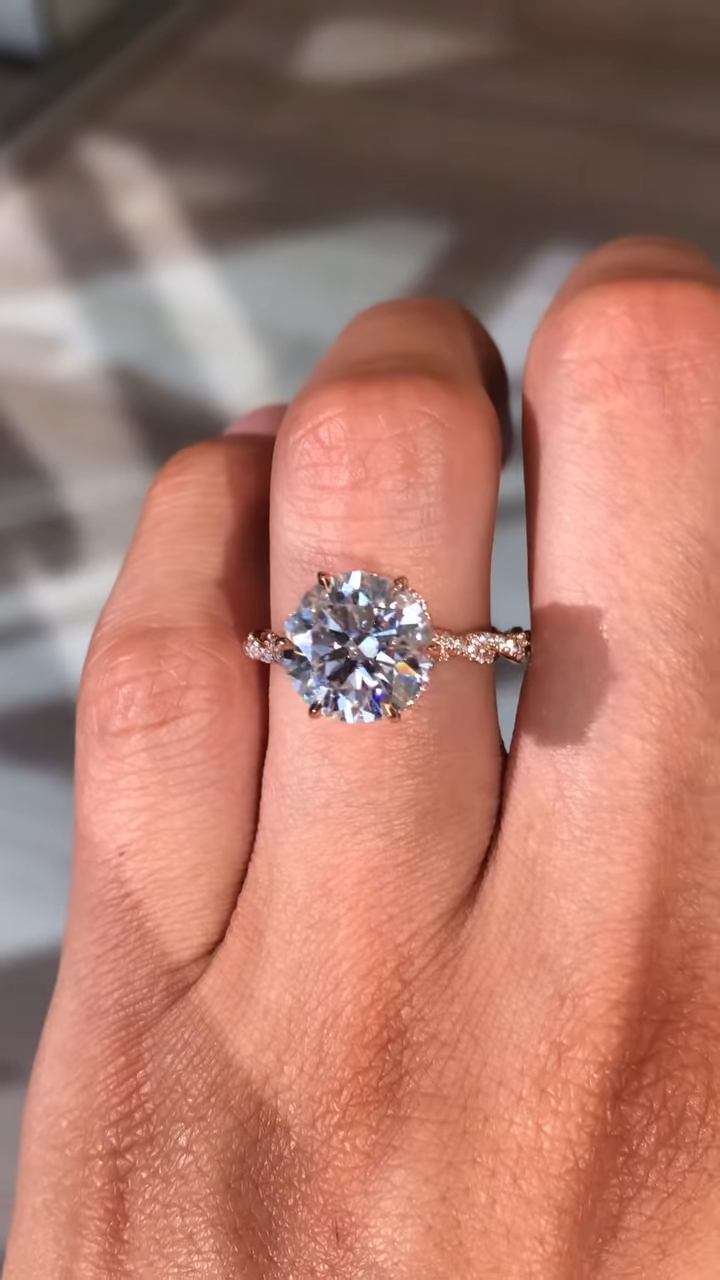 Close-up top view of a round solitaire engagement ring worn on the finger in natural sunlight. The centerpiece features a brilliant round lab grown diamond secured in a classic prong setting, designed to maximize sparkle and light reflection. A delicate rose gold pavé band adds subtle shimmer along the sides of the finger. This elegant engagement ring is also available with a moissanite diamond, offering a sustainable, ethical alternative with exceptional fire and brilliance.