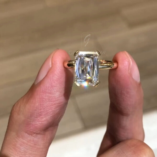 A stunning close-up front view of a solitaire engagement ring featuring a massive elongated radiant-cut center stone. The gem, likely a premium lab grown diamond or high-quality Moissanite, is oriented vertically to elongate the finger. It is securely held by double claw prongs in 18k yellow gold. The stone's "crushed ice" faceting pattern creates a mesmerizing effect, reflecting light in every direction. This sustainable luxury piece blends modern architectural design with the timeless brilliance of a large, colorless diamond alternative.