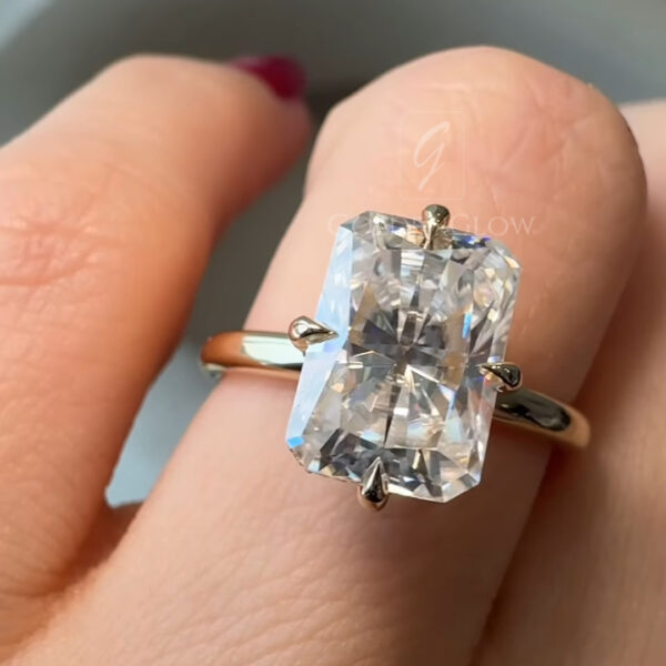 Angled top view of a radiant cut diamond ring resting on a finger, featuring a high-set four-prong yellow gold setting. The elongated diamond sparkles under soft ambient lighting, revealing detailed facet patterns and clean edges. The polished gold band curves smoothly around the finger, offering a timeless minimalist look. Burgundy nail polish and a neutral background create contrast that draws attention to the center stone.