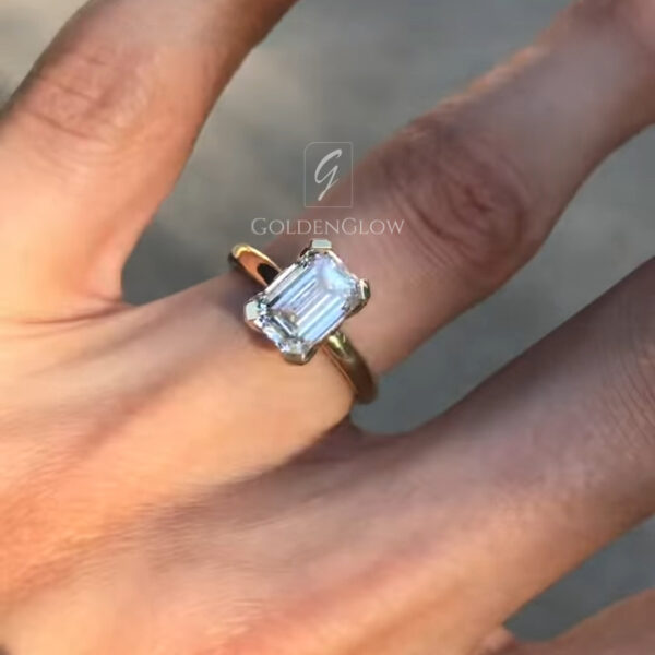 A dynamic angled view of the solitaire ring on a finger, showcasing the height and clarity of the emerald-cut lab grown diamond or Moissanite. The yellow gold prongs clasp the clipped corners of the stone securely. Light reflects off the large table and step facets, producing distinct flashes of white light typical of high-quality step cuts. This perspective emphasizes the clean lines of the Art Deco-inspired design and the warmth of the gold metal against the skin.