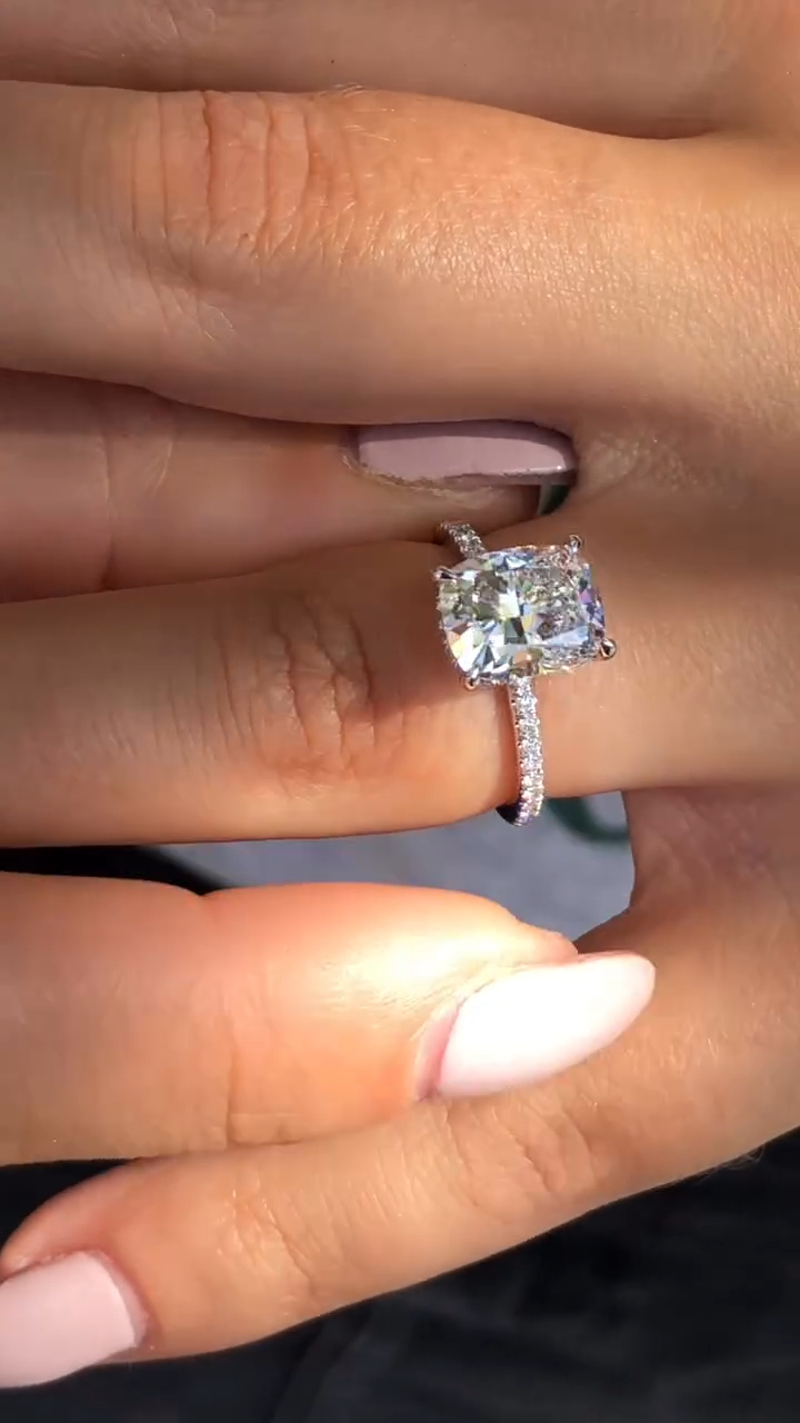 A glamorous side-angle shot of an elongated cushion-cut engagement ring worn on a hand with pink nails. The 18k white gold or platinum band is lined with micro-pavé lab grown diamonds, adding a continuous line of sparkle. The center stone sits high in a setting with a "hidden halo," allowing light to enter from the sides. This lifestyle image highlights the ring's impressive height and the delicate craftsmanship of the pavé work, perfect for a bride who loves romantic detailing.