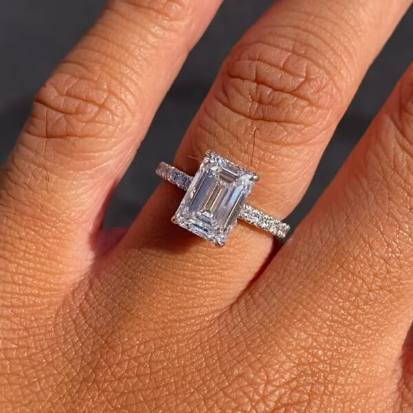 Top view of an emerald-cut solitaire engagement ring worn on the finger in natural sunlight. The step-cut diamond displays crisp lines and mirror-like brilliance, emphasizing its elegant rectangular shape. A pavé-set band adds refined sparkle while keeping the design timeless and balanced. Crafted for modern sophistication, this emerald engagement ring is also available with a moissanite diamond option, offering a sustainable alternative with exceptional clarity and fire.