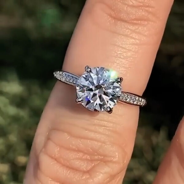 Front-facing close-up of a round engagement ring worn on the finger outdoors, featuring a brilliant diamond secured in a four-prong setting. The slim band is accented with small round stones that add subtle sparkle along the shoulders. Natural sunlight highlights crisp facets and bright reflections. This image allows visual comparison between the clean brilliance of a lab grown diamond and the intense fire often associated with a moissanite diamond.
