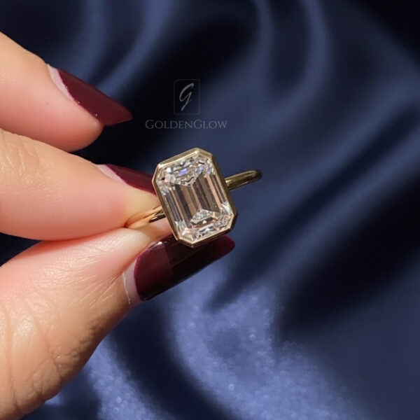 Close-up of a yellow gold ring held between fingertips against a deep blue satin background. The ring features an emerald-cut center stone in a sleek bezel setting with crisp step facets reflecting bright white light. The elongated shape highlights clean geometry and refined craftsmanship. This image compares the visual elegance of a lab grown diamond and a moissanite diamond, emphasizing clarity, sparkle, and modern minimalist design suitable for an engagement ring.