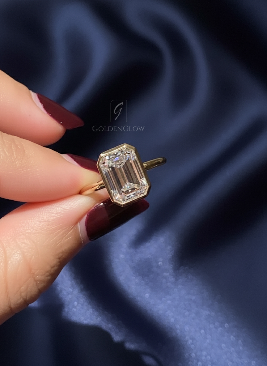 Close-up of a yellow gold ring held between fingertips against a deep blue satin background. The ring features an emerald-cut center stone in a sleek bezel setting with crisp step facets reflecting bright white light. The elongated shape highlights clean geometry and refined craftsmanship. This image compares the visual elegance of a lab grown diamond and a moissanite diamond, emphasizing clarity, sparkle, and modern minimalist design suitable for an engagement ring.