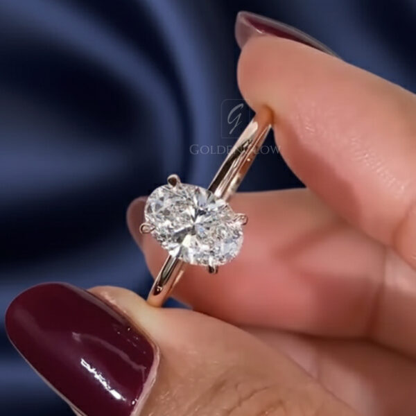Three-quarter close-up of an oval moissanite diamond ring held between fingers, highlighting the stone’s shape and dynamic sparkle. The moissanite is set in a slim yellow gold four-prong solitaire that enhances its size and brilliance. Light creates vivid rainbow flashes across the facets. A dark blue satin background and rich burgundy nail polish frame the ring, emphasizing the bold fire and clarity of the moissanite diamond.
