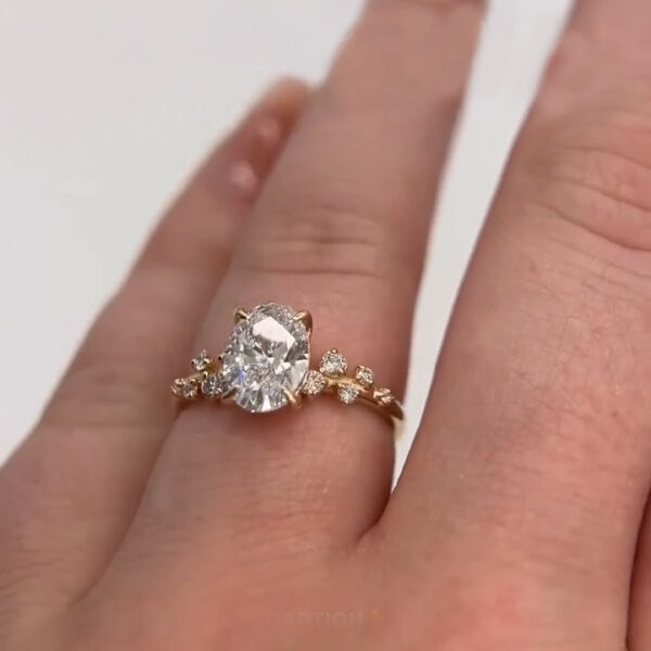 Front-facing view of a diamond oval engagement ring worn on the finger, highlighting the floral-style side accents and polished yellow gold band. The center stone reflects bright flashes of light, offering brilliance comparable to a moissanite diamond. This image captures the ring’s romantic elegance, sustainable gemstone choice, and fine detailing, making it perfect for showcasing modern engagement jewelry with charm.