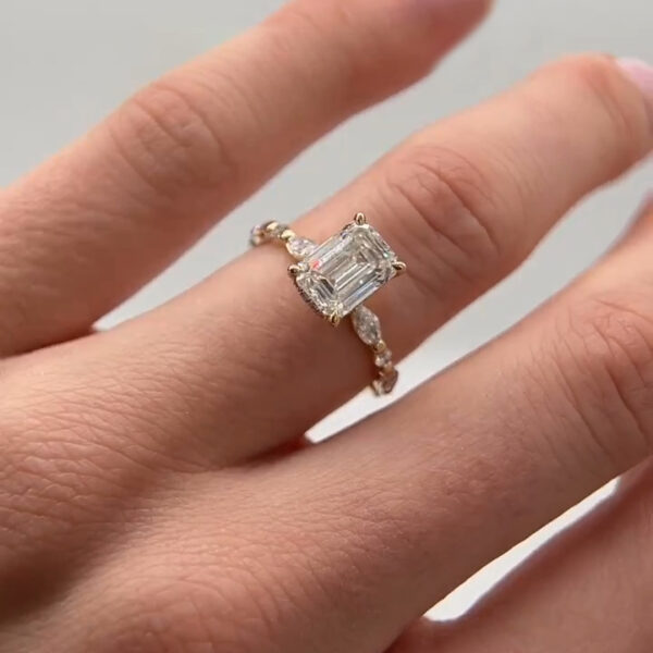 Moissanite diamond engagement ring displayed on a hand against a clean neutral background. The ring showcases an emerald cut moissanite diamond in a yellow gold prong setting, accented by marquise and round stones along the band. The step-cut facets reflect bright flashes of light, emphasizing clarity and symmetry. The slim gold band complements the elongated center stone, highlighting elegant design, sparkle, and contemporary craftsmanship.