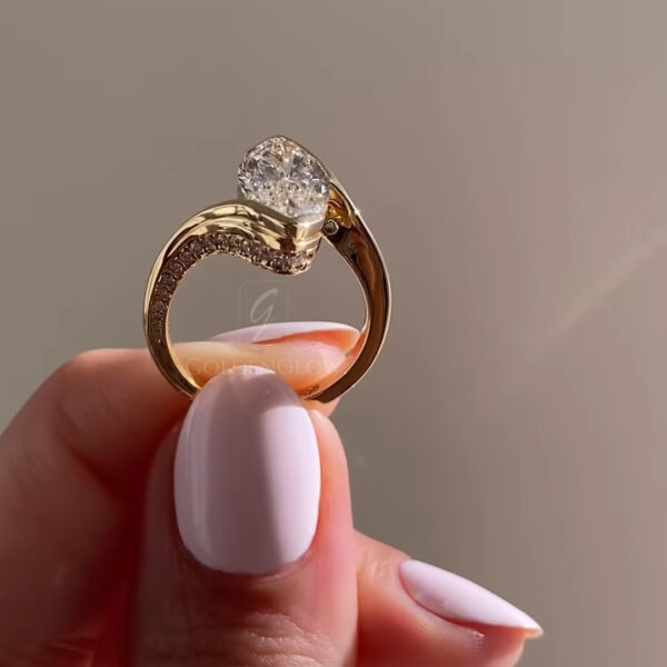 Back view of the yellow gold ring shown upright between fingertips, displaying the open underside of the setting. The oval-cut diamond sits securely in a raised mount, allowing light to pass through the stone. Pavé diamonds line one side of the band, following a graceful, asymmetric curve. Soft natural lighting enhances the contrast between the smooth polished gold and the shimmering accent stones against a minimal beige background.