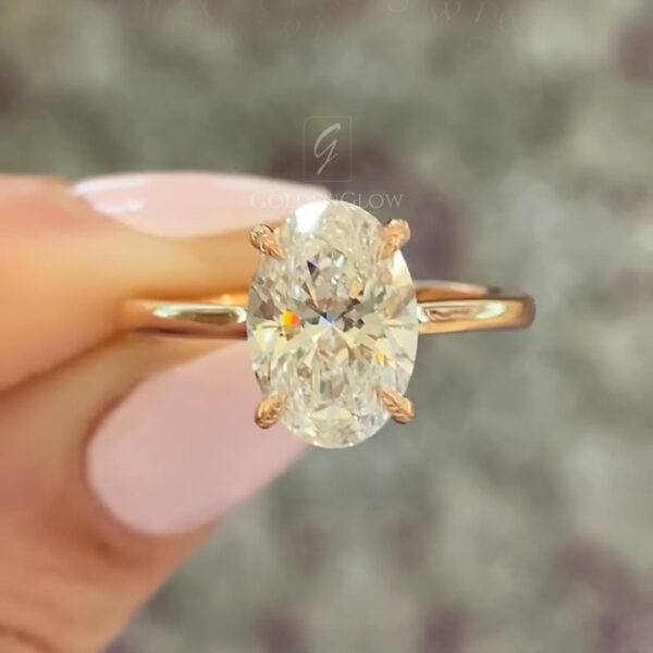 Close-up side profile of a diamond or moissanite engagement ring in polished yellow gold, held between manicured fingers. The oval-cut center stone sits in a refined basket setting with petite pavé accent diamonds beneath the gallery, adding subtle sparkle from the side view. The slim gold band reflects warm light, emphasizing craftsmanship, symmetry, and ethical luxury. Soft brick background enhances the brilliance and clarity of the gemstone.