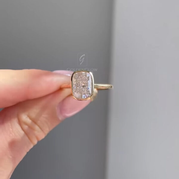 Close-up front view of a rectangular cushion solitaire engagement ring held between manicured fingers. The centerpiece features an elongated cushion lab grown diamond secured in a smooth yellow gold bezel setting that highlights clean lines and modern elegance. The polished gold band adds warmth and simplicity to the design. This ring is also available with a moissanite diamond, offering an ethical, sustainable alternative with exceptional brilliance and fire.