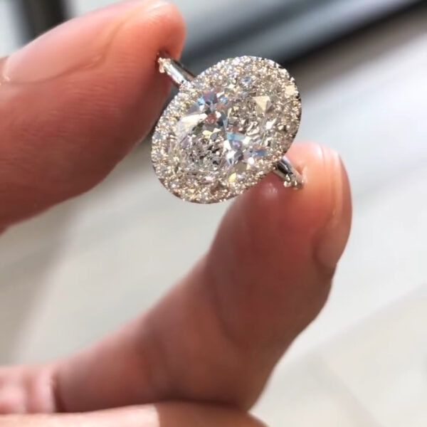 A stunning side profile view of a handcrafted diamond engagement ring held between fingertips. This angle reveals the intricate high-setting architecture, showcasing a massive oval-cut center stone resting on a delicate gallery. The slim band features micro-pavé detailing that adds extra shimmer from every angle. The light passes through the diamond's pavilion, emphasizing its depth and clarity. This view highlights the structural elegance and the hidden details of the basket setting.