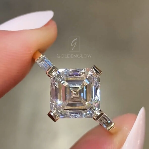 Close-up of an Asscher-cut center stone engagement ring held delicately between two fingers with light pink nails. The square step-cut stone shows crisp concentric facets and strong light return, secured by four claw prongs in a yellow gold setting with tapered baguette side stones. This fine jewelry image highlights a lab grown diamond option while also appealing to shoppers comparing a moissanite diamond for brilliance, geometry, and modern elegance in a luxury engagement ring design.