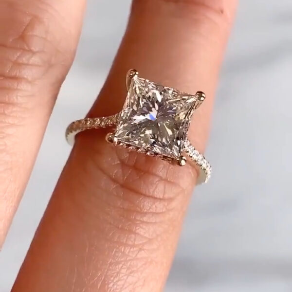 Close-up view of a square-cut engagement ring resting on a finger, showcasing a brilliant diamond secured by four prongs. The pavé band sparkles with small accent stones that enhance the center stone’s fire and clarity. The lighting highlights crisp facets and clean lines, offering a comparison look often associated with a moissanite diamond while emphasizing the refined brilliance and symmetry of a lab grown diamond in a modern solitaire setting.