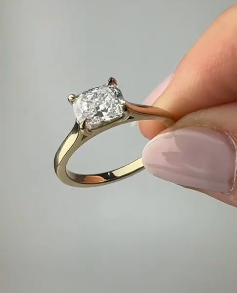 Close-up of a yellow gold solitaire ring held between fingers, featuring a rectangular emerald-cut stone secured by four prongs. The polished band reflects soft studio light while the clear gemstone shows crisp step facets and bright sparkle. This fine jewelry image highlights craftsmanship and clarity, suitable for showcasing a lab grown diamond or a moissanite diamond as an elegant engagement or statement ring from a side profile against a neutral background.