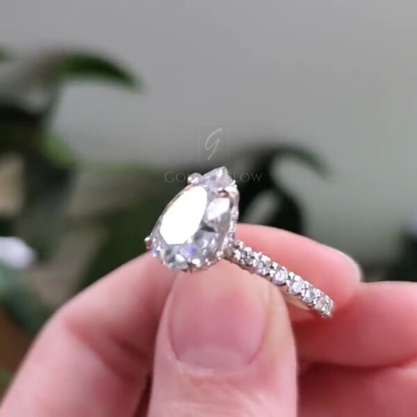 Side profile close-up of a pear-cut clear gemstone ring held between fingers. The image showcases the raised prong setting and the pavé diamonds running along the band’s upper half. Light passes through the gemstone, revealing faceting and clarity. The polished metal band appears sleek and evenly finished. The background is softly out of focus, helping emphasize the ring’s silhouette, height, and detailed workmanship.