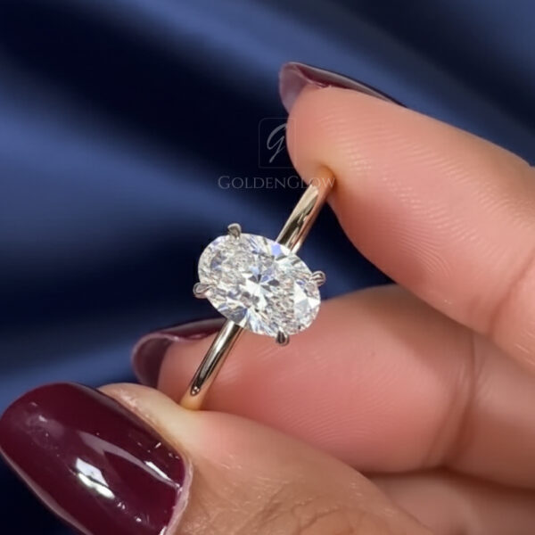 Close-up of a hand wearing an oval moissanite diamond solitaire ring on the ring finger. The moissanite is mounted on a slender yellow gold band with four fine prongs, creating a clean and modern silhouette. The stone shows intense sparkle with bright rainbow flashes under soft lighting. Burgundy manicured nails and a dark blue satin backdrop provide contrast, emphasizing the brilliance and fire characteristic of a moissanite diamond ring. I
