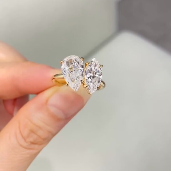 Close-up view of a yellow gold toi et moi engagement ring held between fingers, featuring two elongated diamonds set side by side. One diamond is pear-shaped and the other marquise-cut, each secured with delicate gold prongs. The stones catch the light with sharp brilliance and fire. The polished gold band curves smoothly around the finger area, while a soft, neutral showroom background keeps focus on the contrasting diamond shapes and elegant asymmetrical design.