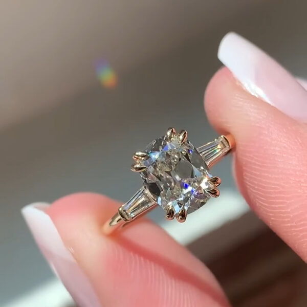 Close-up photograph of a gold engagement ring held delicately between two fingertips with manicured white nails. The ring features a rectangular radiant-cut diamond set vertically in a yellow-gold band. Four prominent claw prongs secure the center stone, which reflects light with sharp, mirror-like facets. Tapered baguette diamonds are set horizontally on each side of the center stone, adding a balanced, elegant look. Soft natural light creates subtle rainbow reflections against a blurred neutral background.
