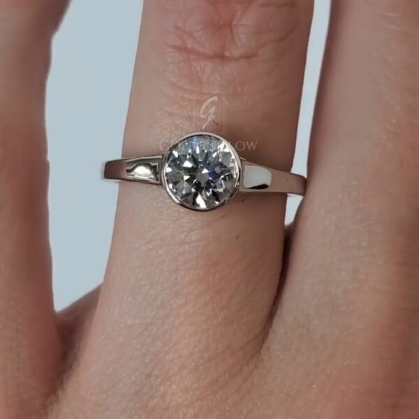 Close-up view of a minimalist solitaire engagement ring worn on a finger, featuring a round brilliant-cut clear gemstone bezel-set in polished white metal. The smooth, slightly tapered band reflects soft studio lighting, highlighting the stone’s crisp facets and symmetrical sparkle. Fine skin texture and natural creases of the finger are visible, adding realism and scale, while the clean white background keeps full focus on the ring’s modern, understated elegance.